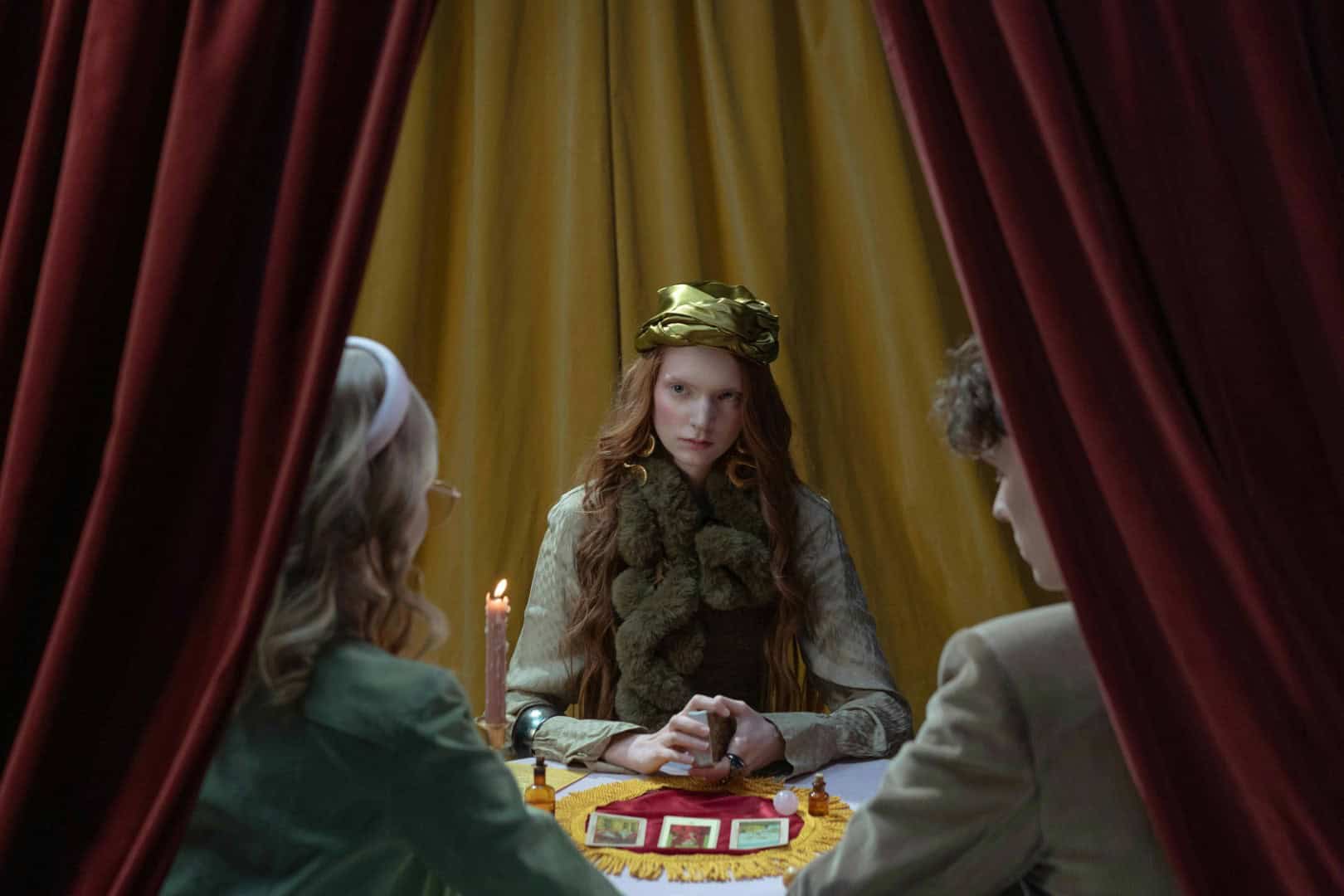 A young woman with red hair wearing a gold turban sits at a table, appearing to lead a tarot reading session in a room with golden curtains.